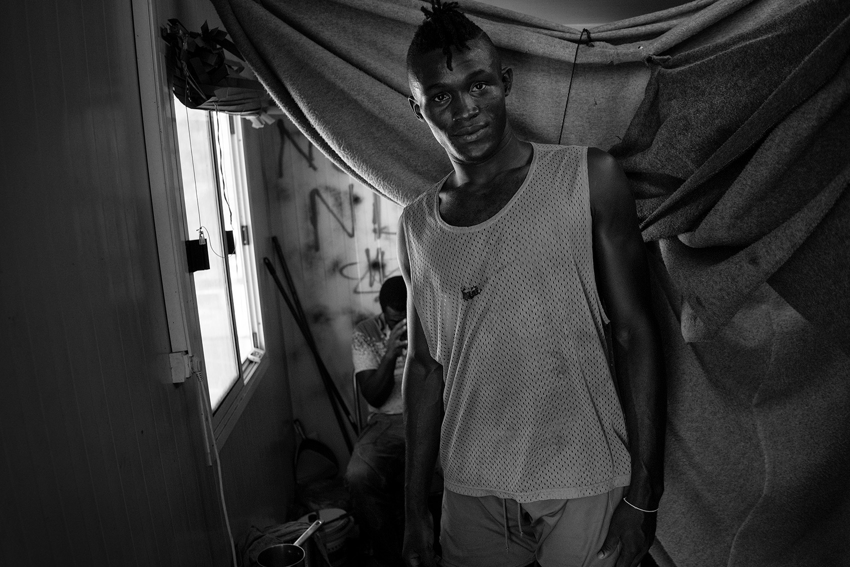 Ali, a professional football player from Guinee, inside his container at the refugee camp Moria. He lives together with 4 other refugees from Africa, their beds divided by blankets, Lesvos, Greece.